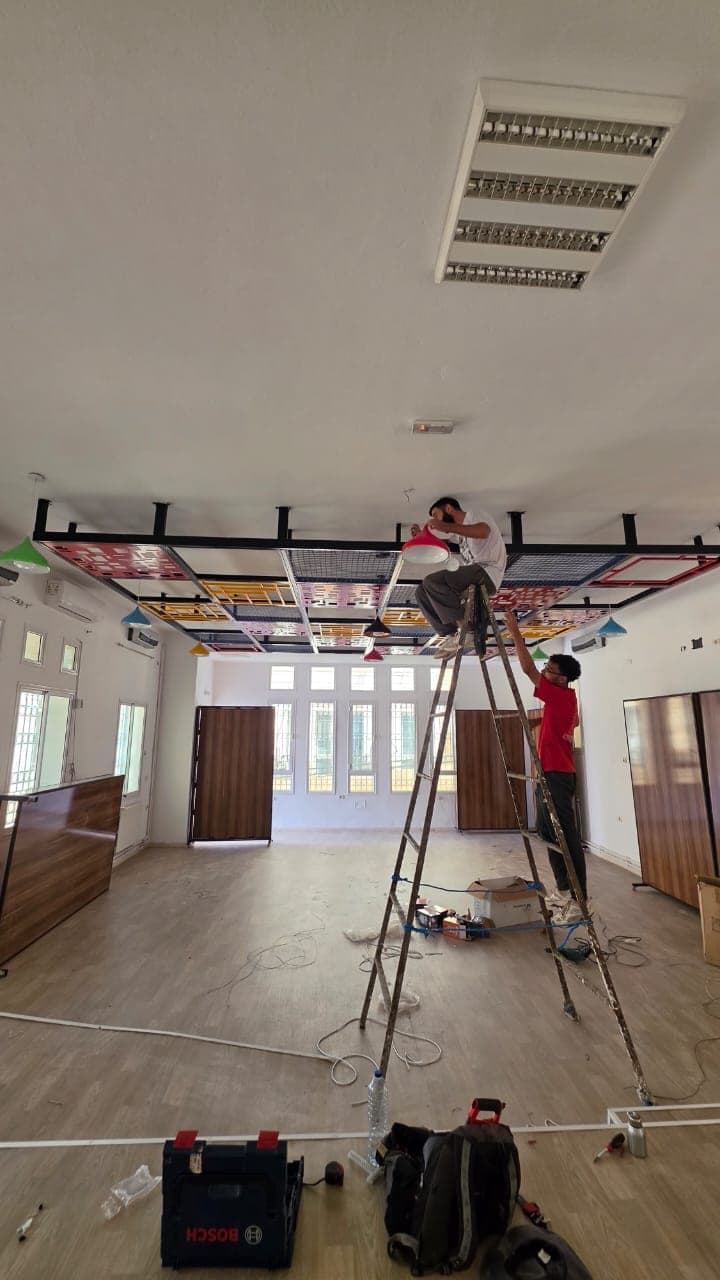 Vue d’ensemble de la salle en chantier : deux personnes posent les suspensions sous le faux plafond coloré, outils et câbles au sol.