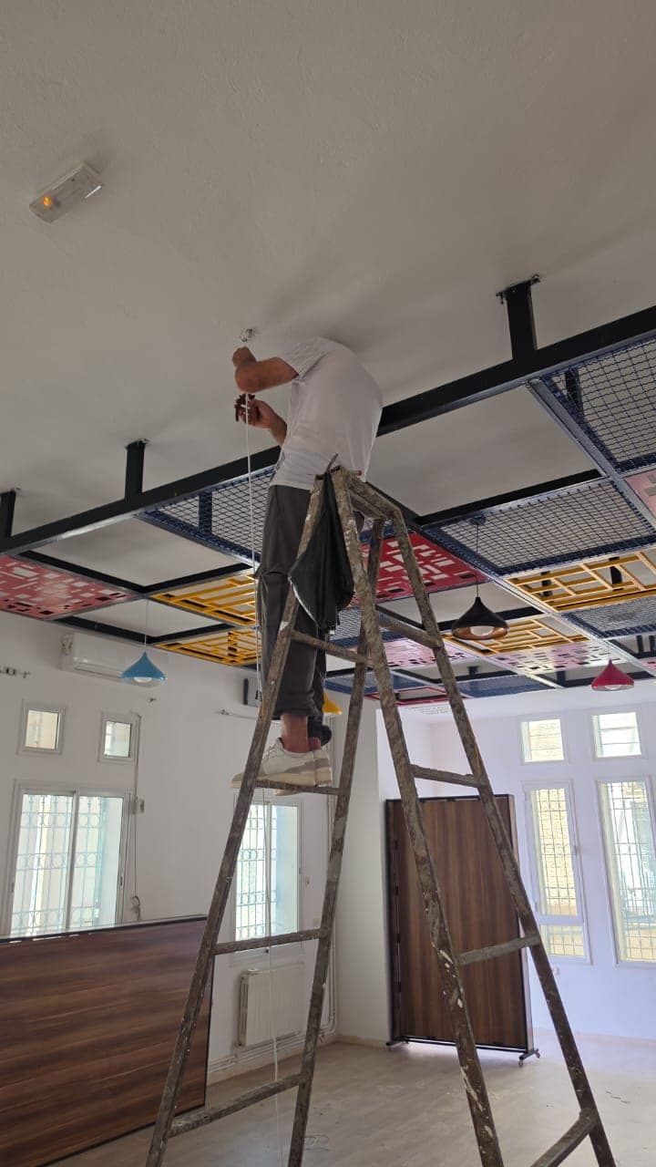 Un ouvrier, sur un escabeau, raccorde un luminaire sous la structure métallique du faux plafond.