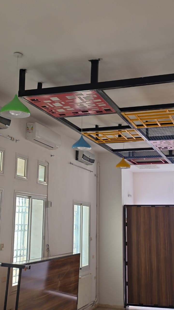 Faux plafond à panneaux ajourés rouges, jaunes et grillagés, avec des suspensions colorées au-dessus de la salle.