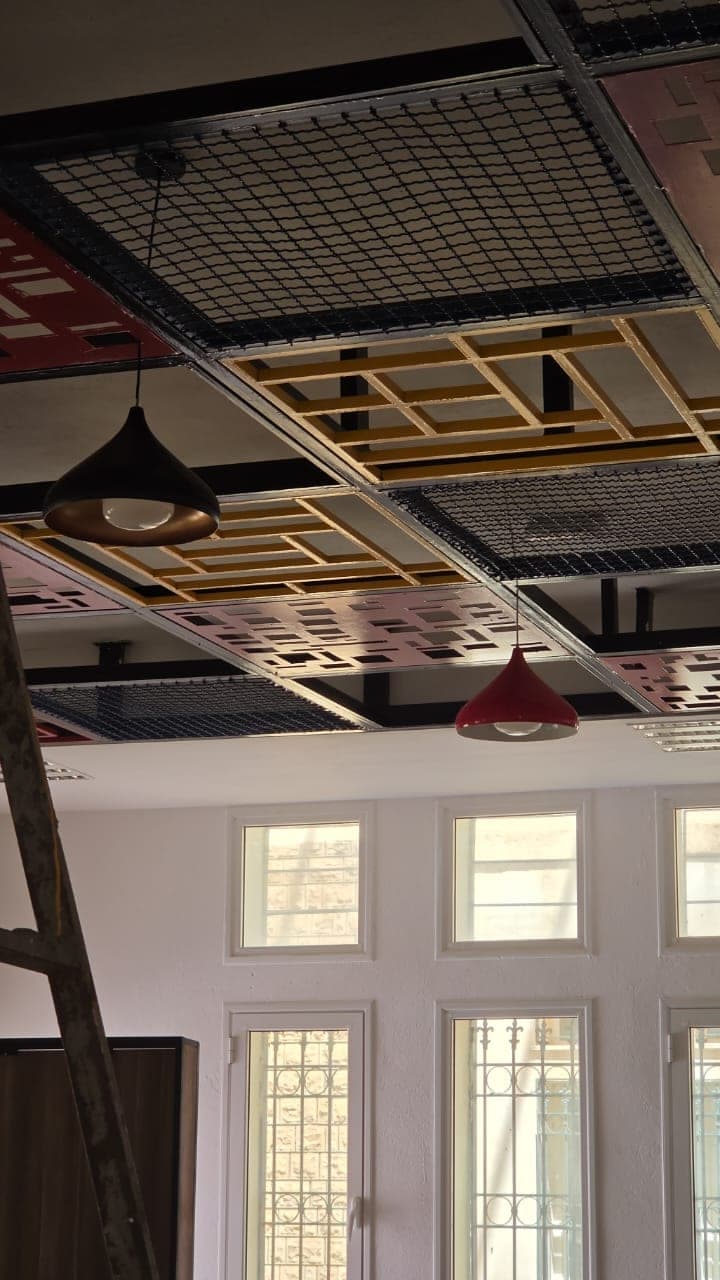 Détail du faux plafond — panneaux ajourés, grilles et cadres jaunes — au-dessus d’une rangée de fenêtres.