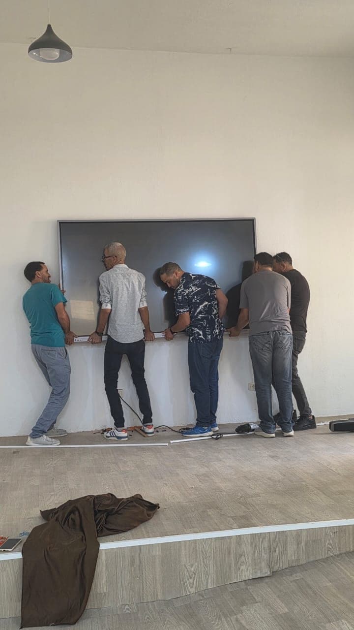 Cinq personnes fixent ensemble un grand écran au mur, sur l’estrade de la salle.