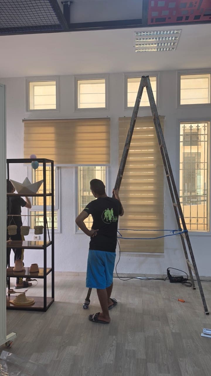 Pose des stores devant les grandes fenêtres ; une étagère d’objets artisanaux se trouve à gauche.