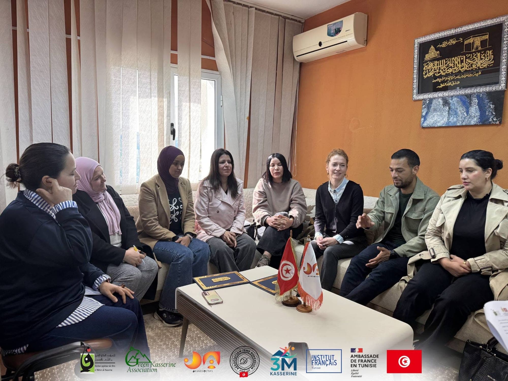 Huit personnes en discussion, assises sur des canapés autour d’une table basse portant un drapeau tunisien et le fanion de l’association.