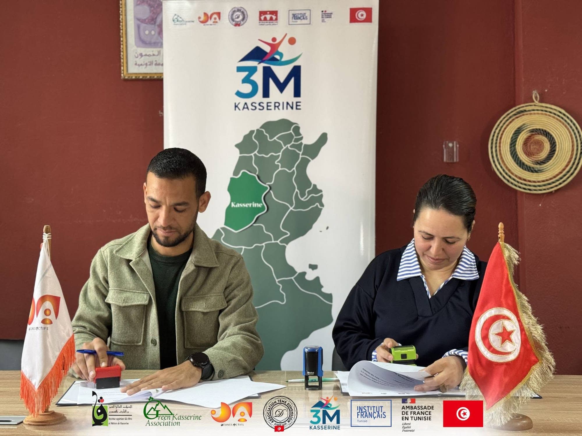 Deux représentants signent et tamponnent des documents, assis devant le kakémono du programme 3M Kasserine.